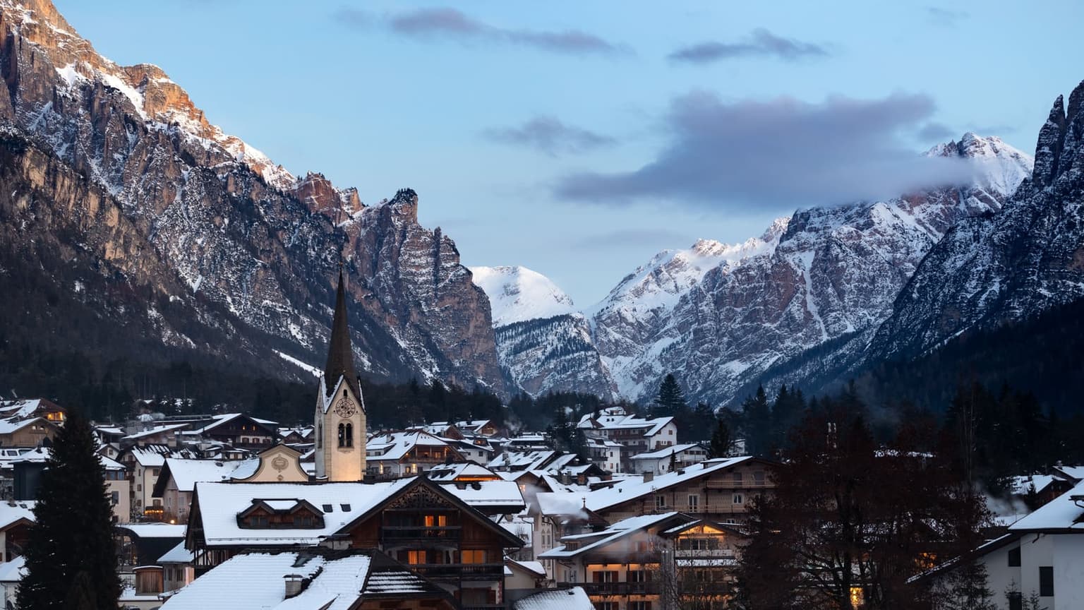 A snow-covered Dolomites village at blue hour, its chalets lit from inside and a church clock tower rising among the roofs, with snowy limestone peaks above the valley