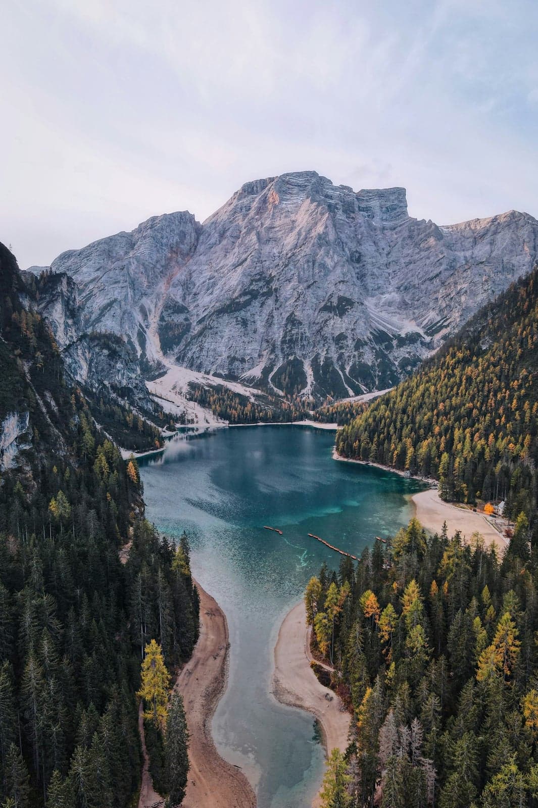 Aerial view of Lago di Braies filling its dead-end alpine valley, turquoise water framed by dense pine and larch forest, a bare gray Dolomite peak rising directly beyond the lake, with the wooden boathouse and its rowboat dock visible in miniature on the near shore