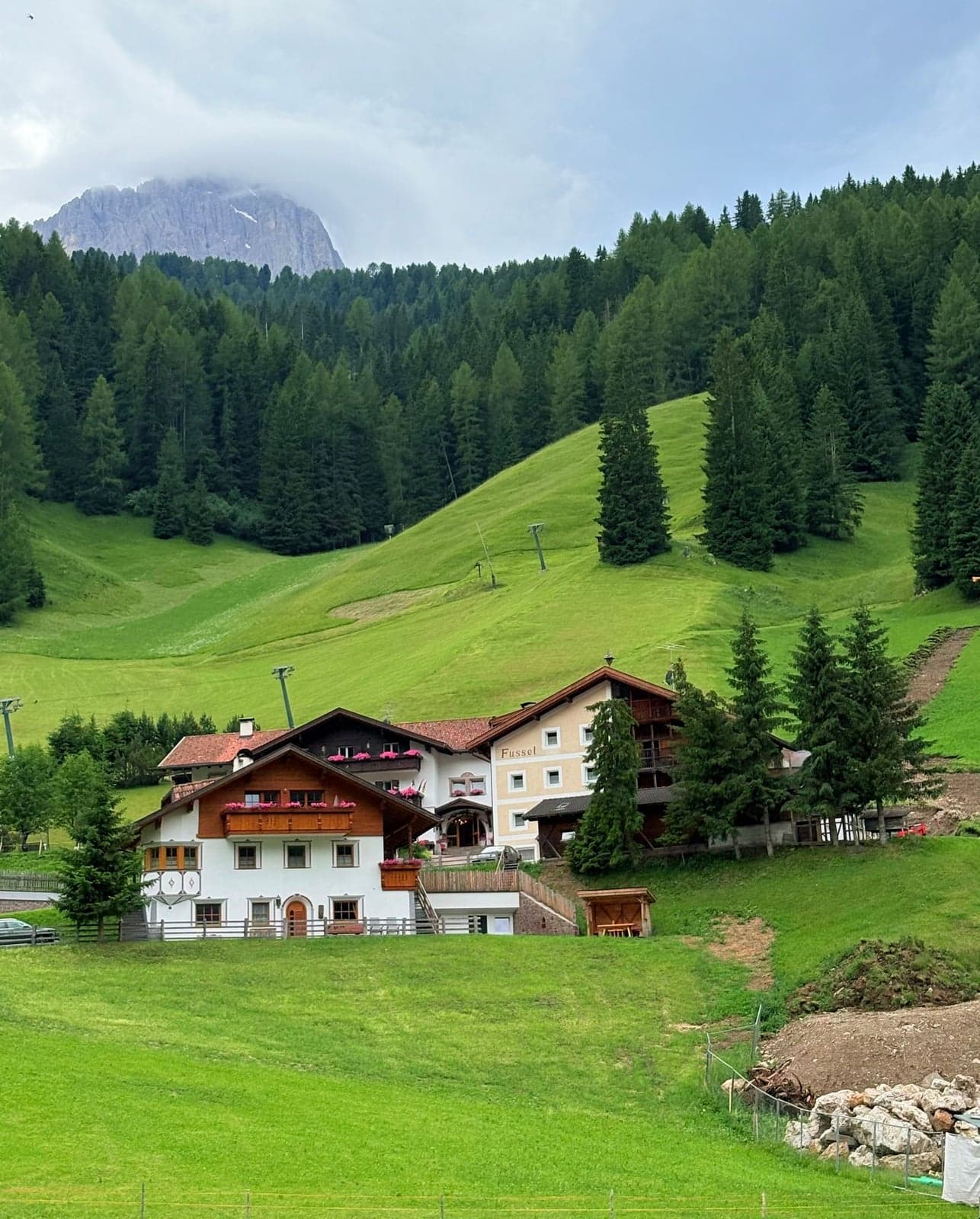 A traditional South Tyrol guesthouse in an alpine valley, white stucco walls, dark wood balconies spilling over with pink geraniums, an arched wooden entrance, set against a green hillside, pine forest, and a rugged, partly cloud-wrapped Dolomite rock face