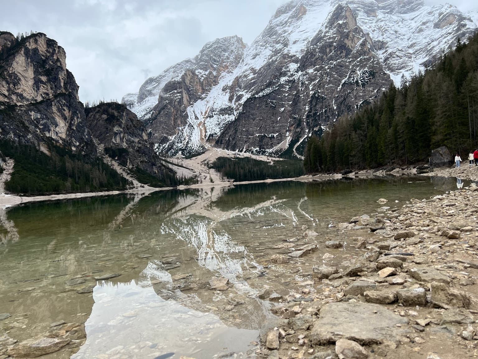 Lago di Braies under an overcast shoulder-season sky, low water exposing the rocky shoreline and snow still clinging to the peaks