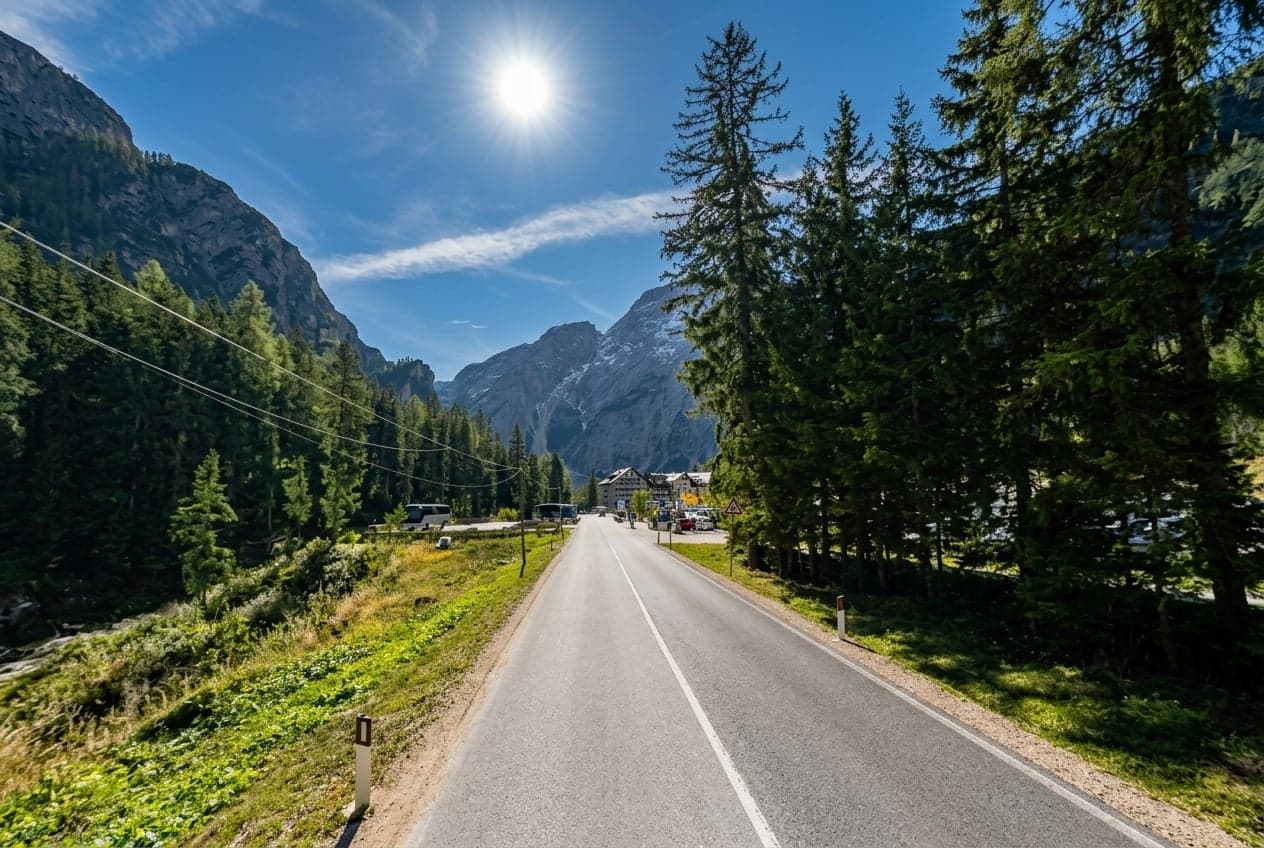 The single-lane road climbing into the Pragser Tal valley toward Lago di Braies, a dashed white centerline, dense pine forest on both sides, a jagged Dolomite rock wall to the left and a snow-streaked peak ahead, the hotel and parking cluster visible at the base of the slope