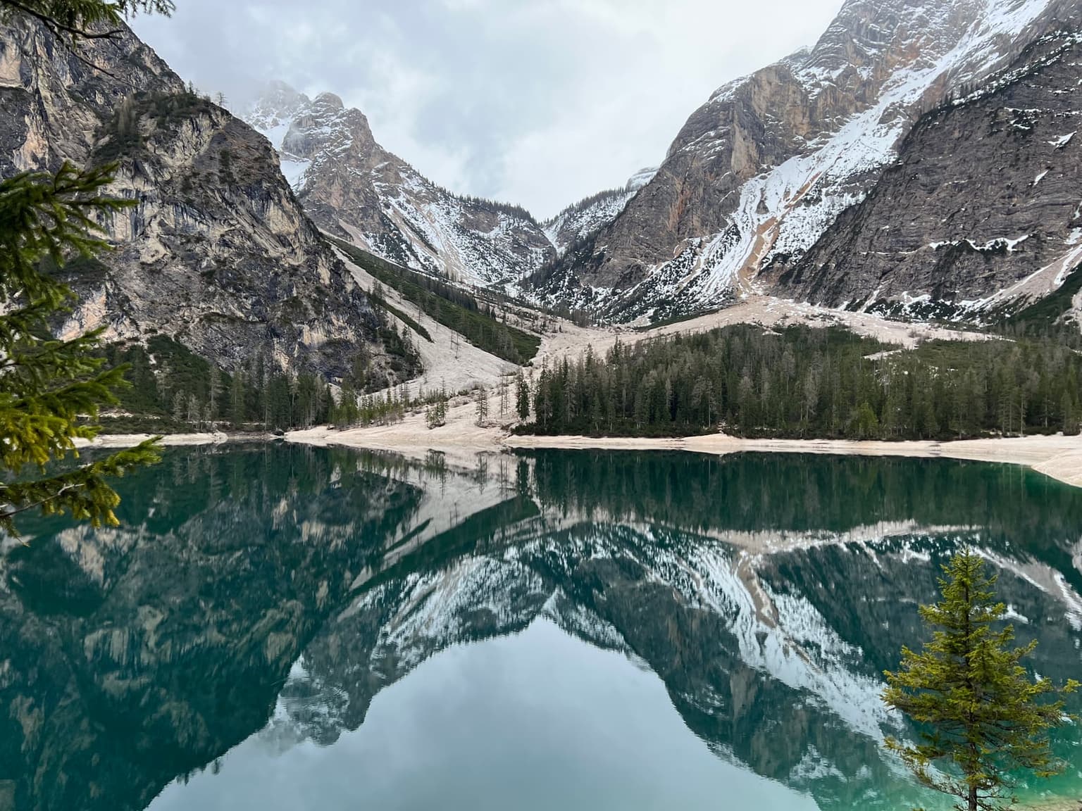 A near-empty Lago di Braies in moody shoulder-season light, the snow-dusted peak mirrored in glassy water at the head of the valley, seen through pine branches
