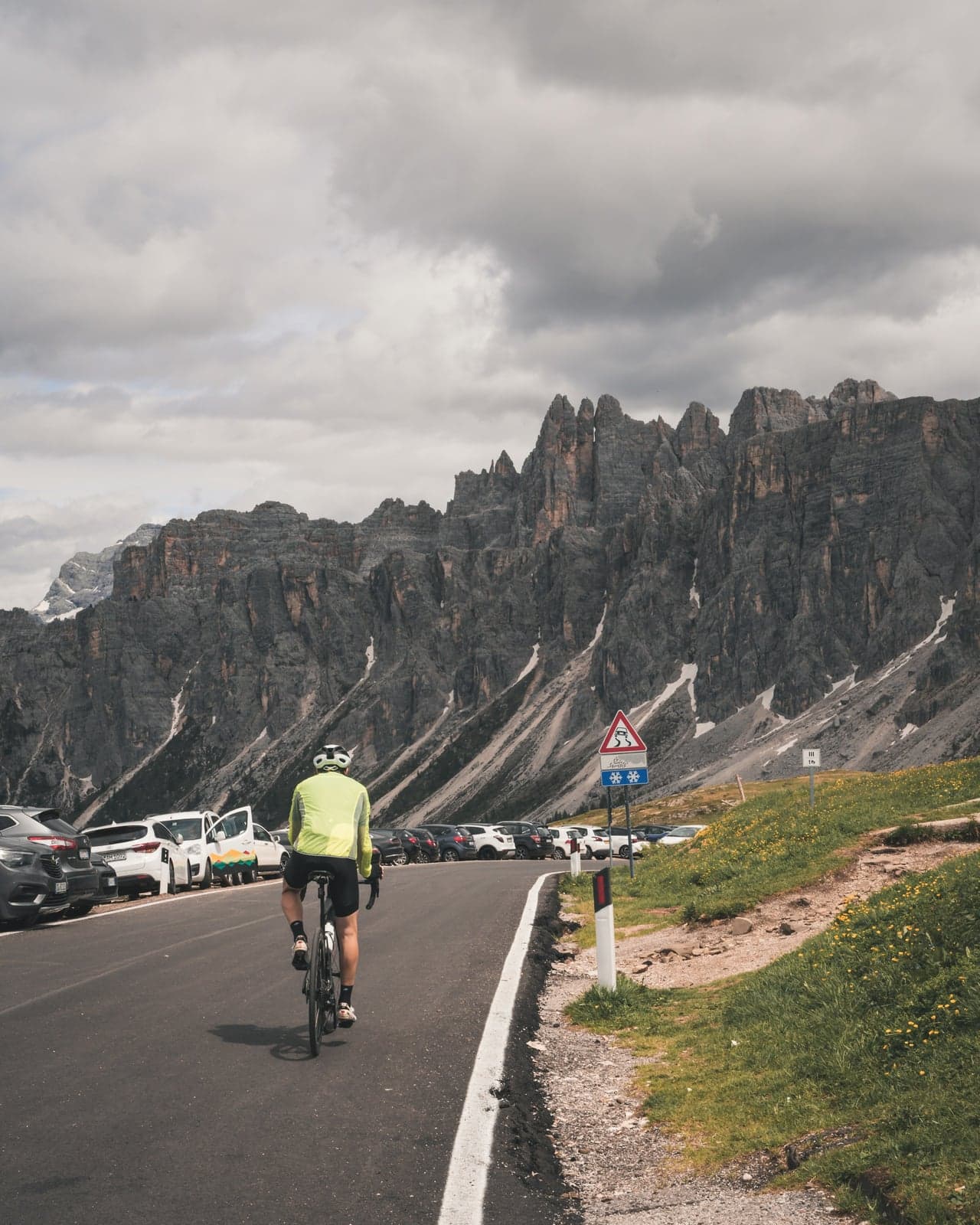 A road cyclist climbing a mountain road past a long row of parked cars, a jagged gray rock wall behind under heavy clouds