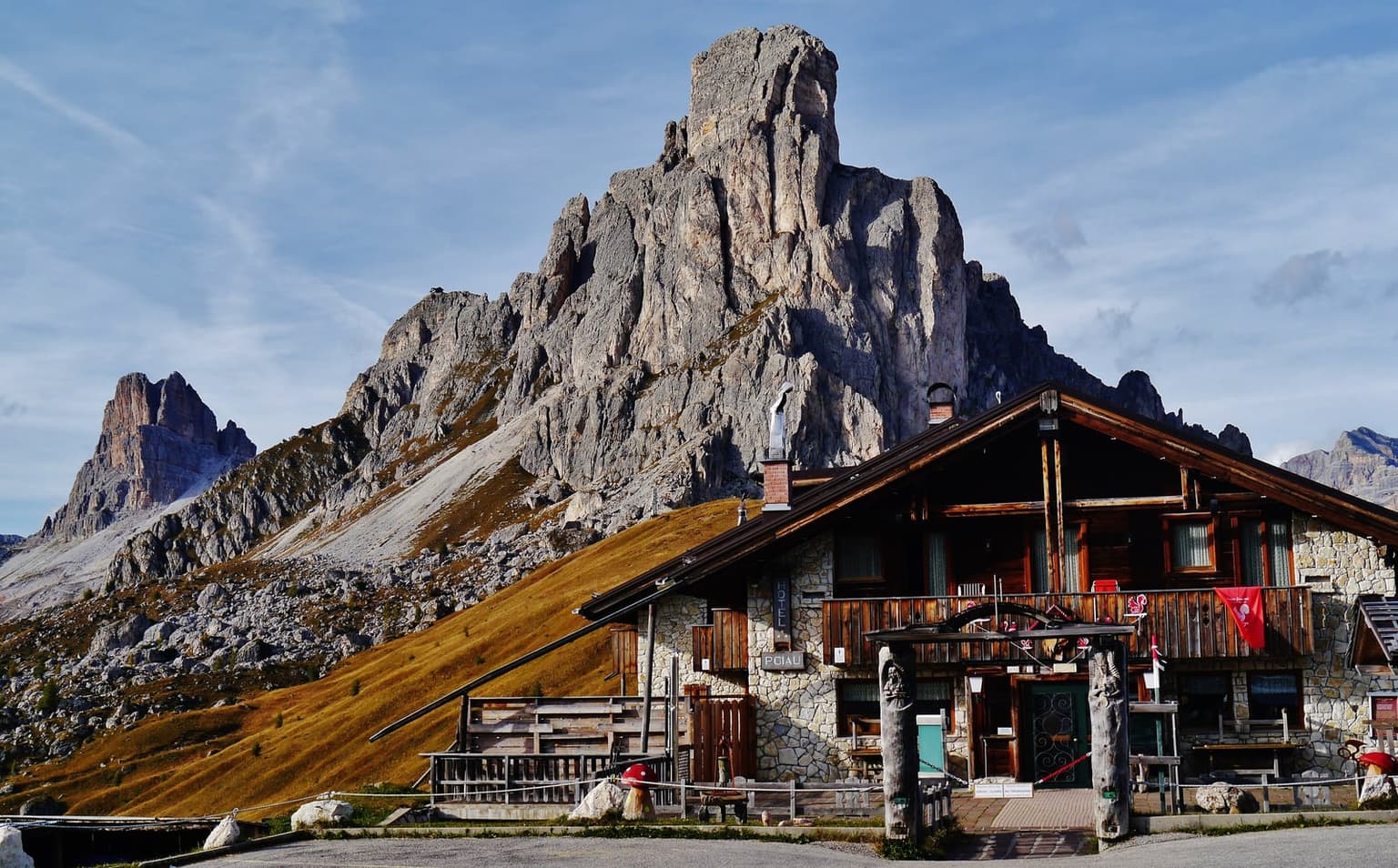 The Hotel Ristorante Passo Giau, a stone and dark timber chalet with a long wooden balcony, Ra Gusela rising right behind it