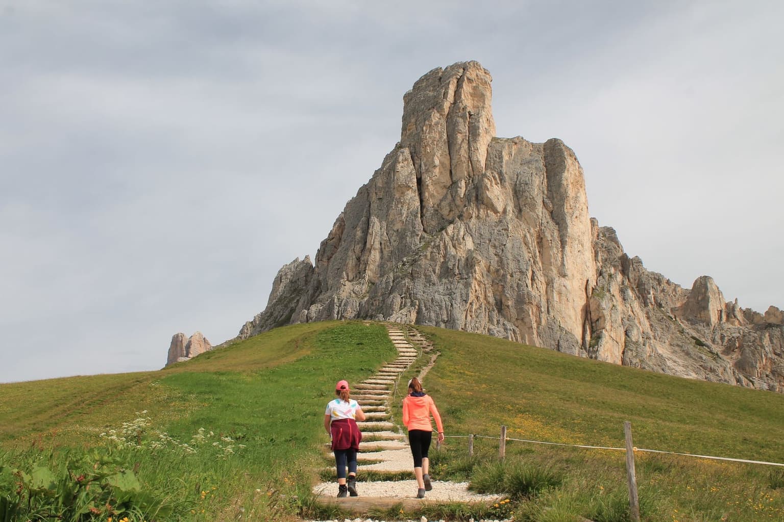 Two walkers on a stepped stone path up the grassy slope toward the rock tower of Ra Gusela, a wooden fence along the right