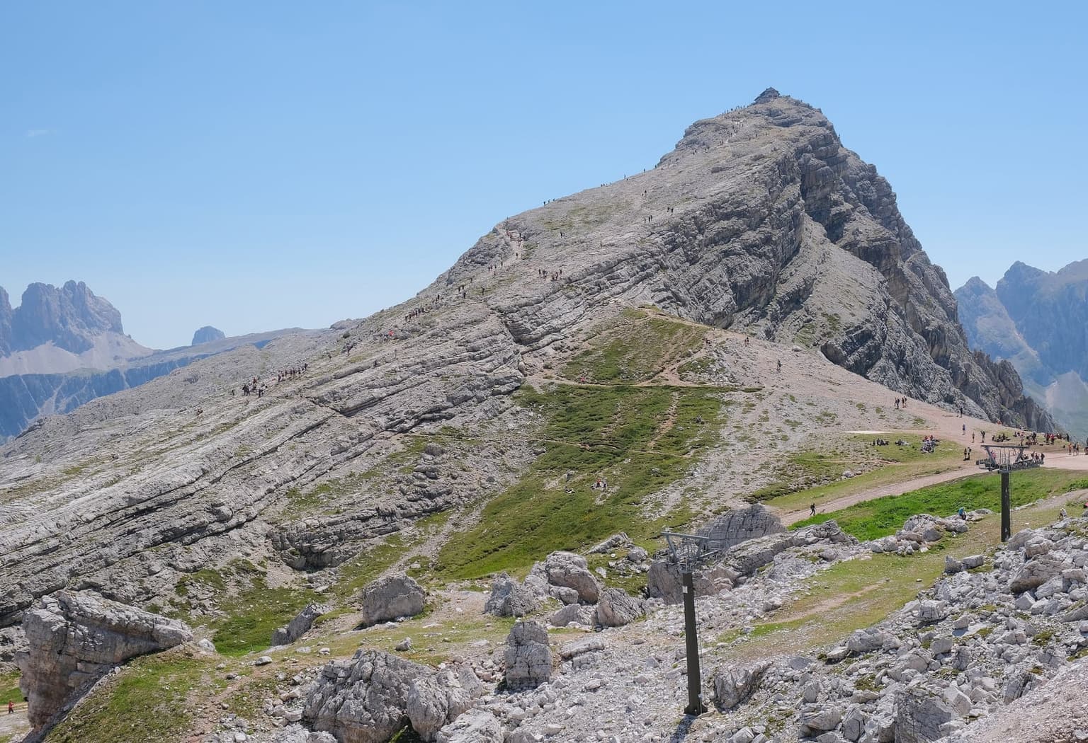 Monte Nuvolau, its bare gray limestone slabs tilting up to Rifugio Nuvolau on the summit, lines of hikers on the rock, and chairlift pylons and walkers resting on a grassy saddle in front