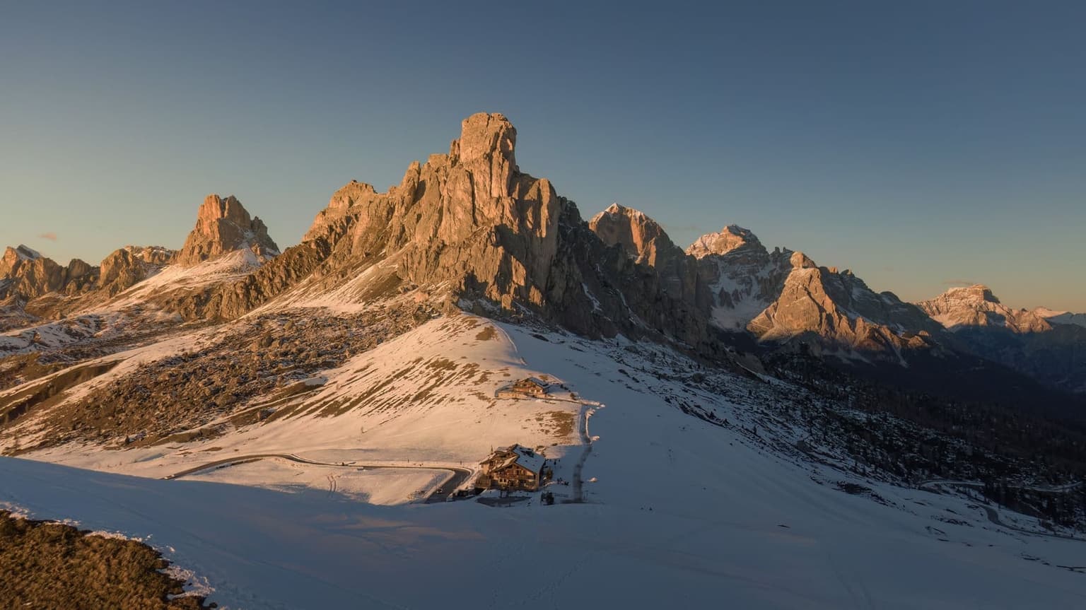Passo Giau under snow, Ra Gusela and the peaks beside it lit warm by a low sun, the hotel and a plowed road crossing the snowfield