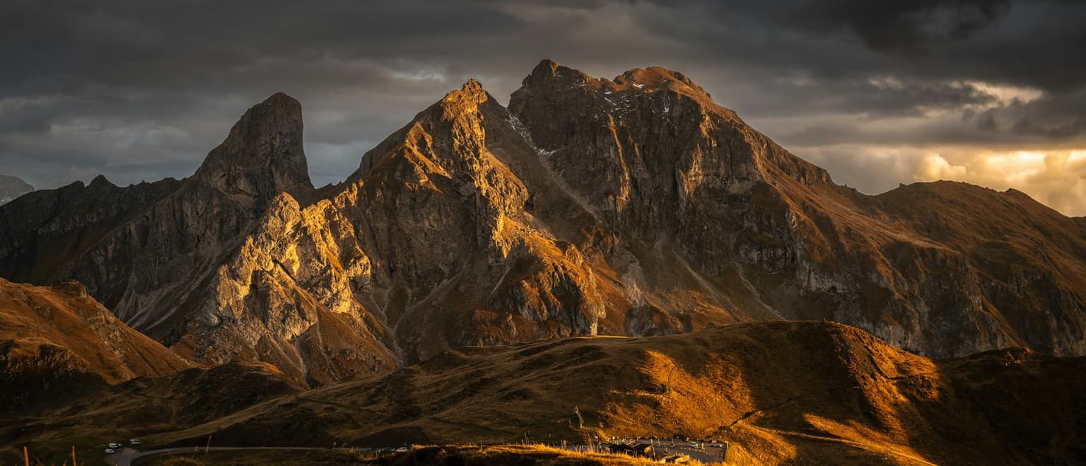 Torre Dusso and Monte Cernera lit orange by the last sun under dark storm clouds above Passo Giau, the grassy ridges below in shadow, a few cars along the road and the pass buildings at the bottom edge