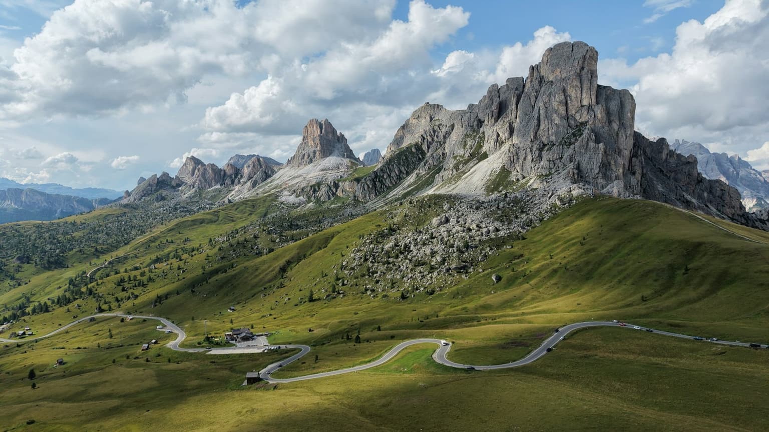 Passo Giau from above: the pale rock wall of Ra Gusela over green pastures, the road winding across the meadow past the small hotel and parking lot at the pass