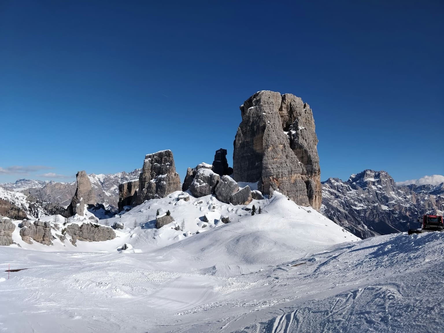 The Cinque Torri rock towers above groomed snow, with a piste groomer at the right edge and Dolomite peaks beyond