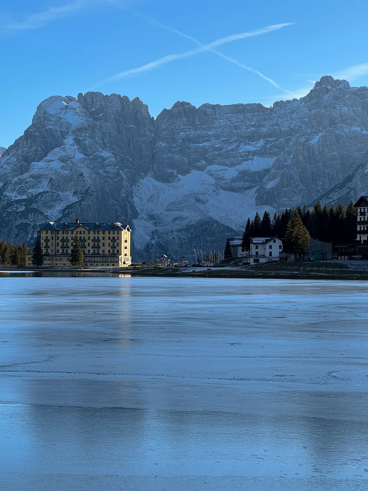 A frozen lake below a large yellow hotel and a steep, snow-dusted Dolomite rock wall