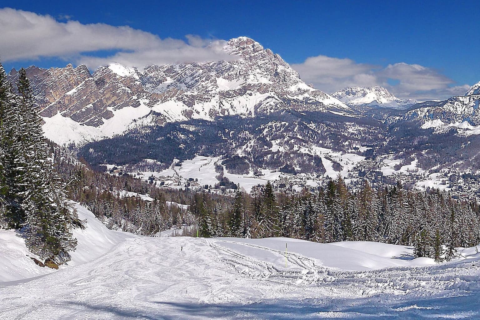 A wide groomed ski slope lined with fir trees, looking over a valley and a town toward a large snowy massif