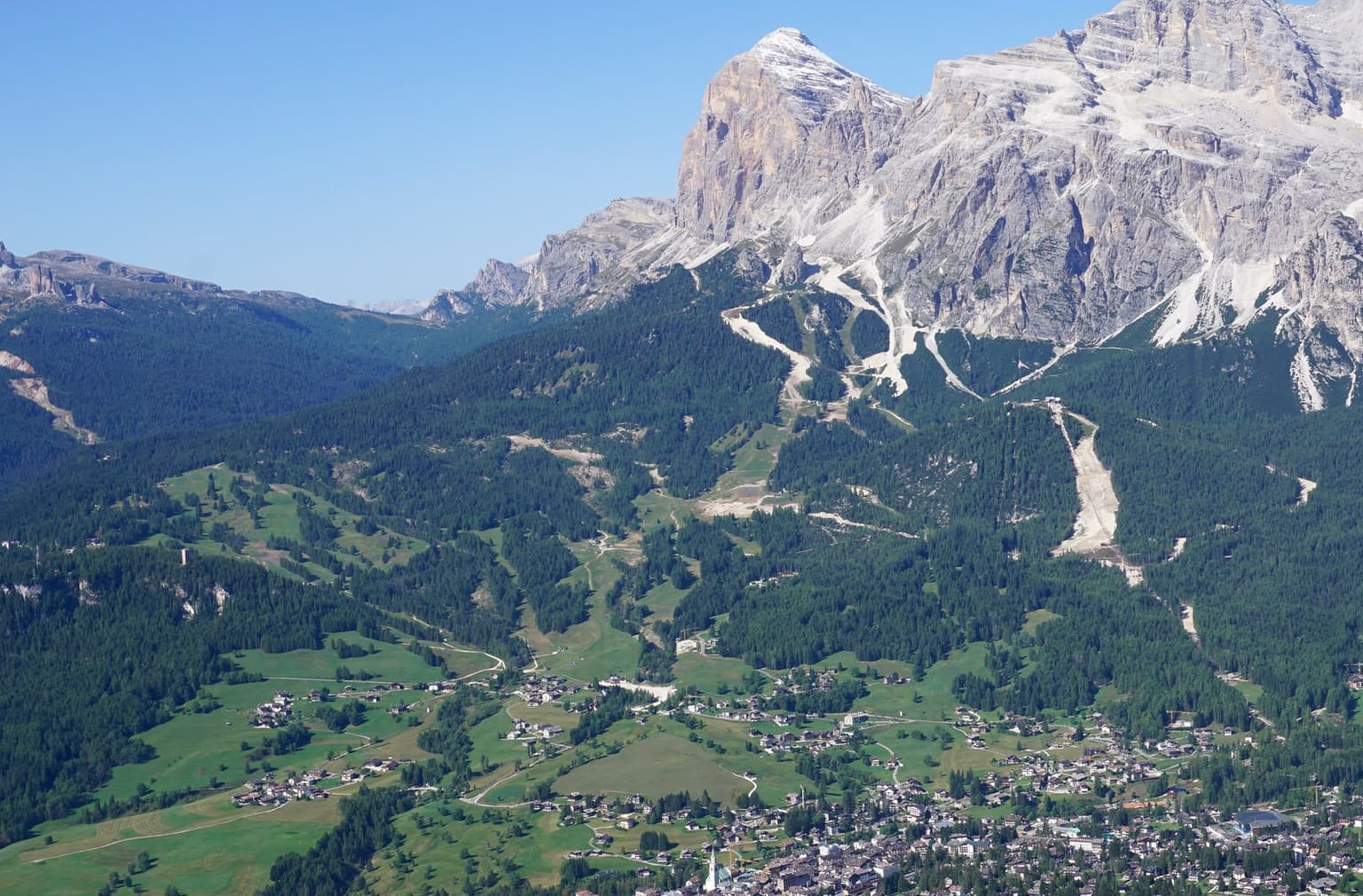 The Olympia delle Tofane course seen from Faloria in summer, a bare strip through the forest below the Tofane, with Cortina at the bottom of the frame