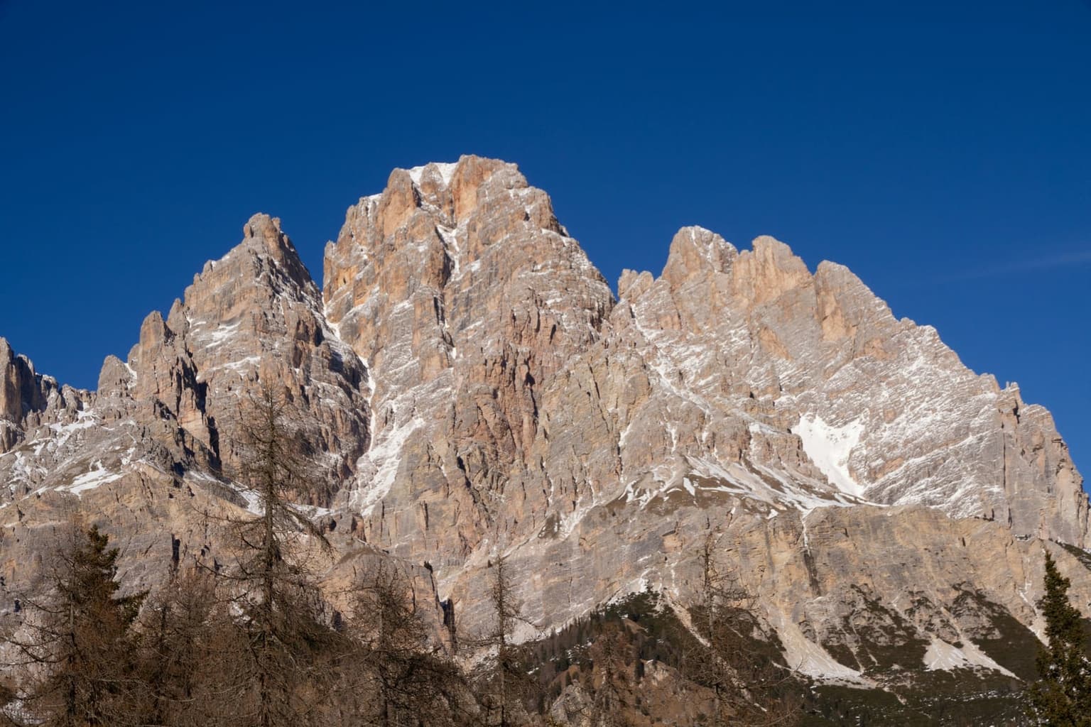 A sunlit Dolomite massif with snow in the gullies and bare trees in the foreground