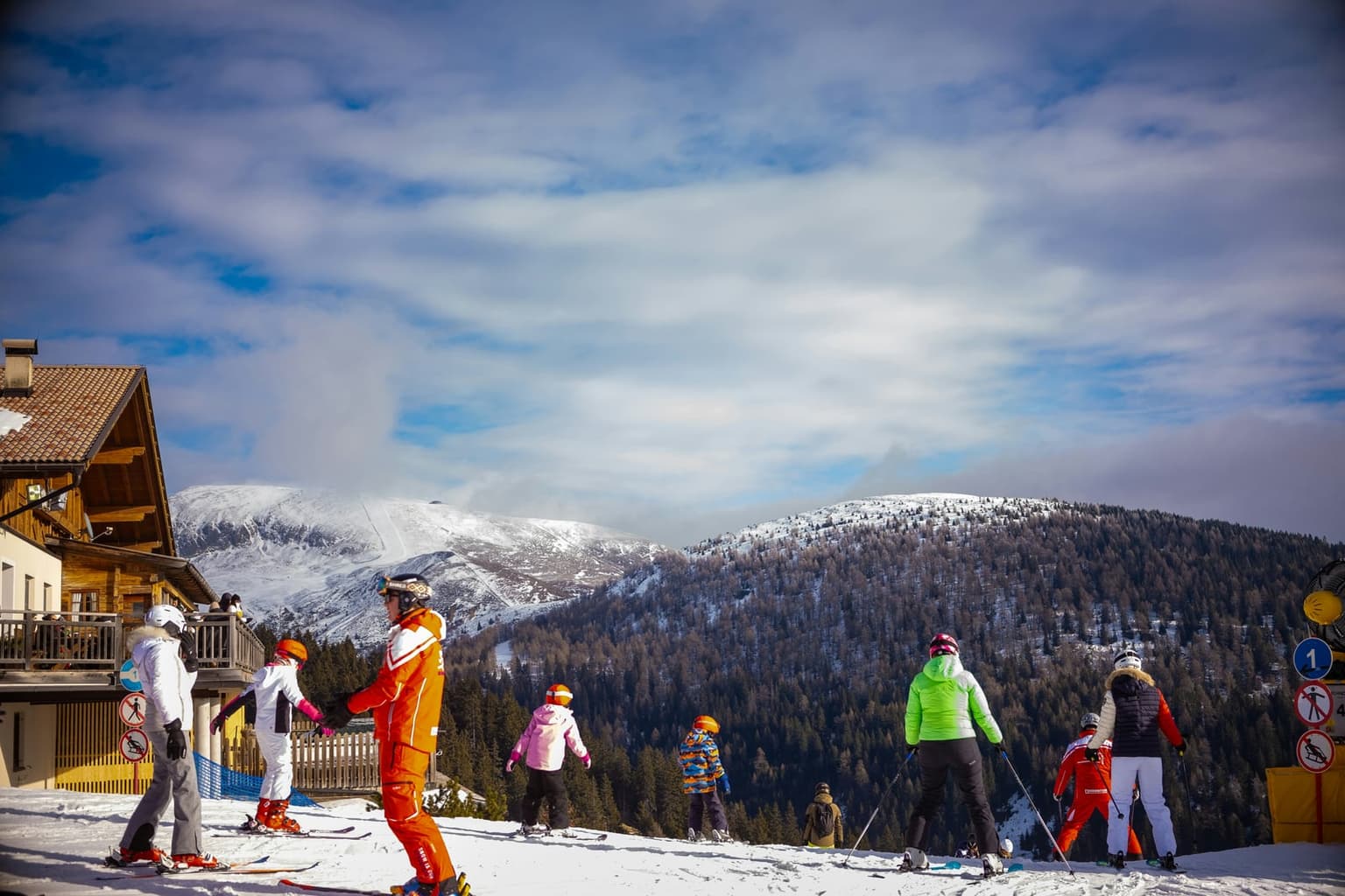 A group of skiers in colorful jackets and helmets, some of them children, gathered on a snowy slope beside a wooden mountain lodge