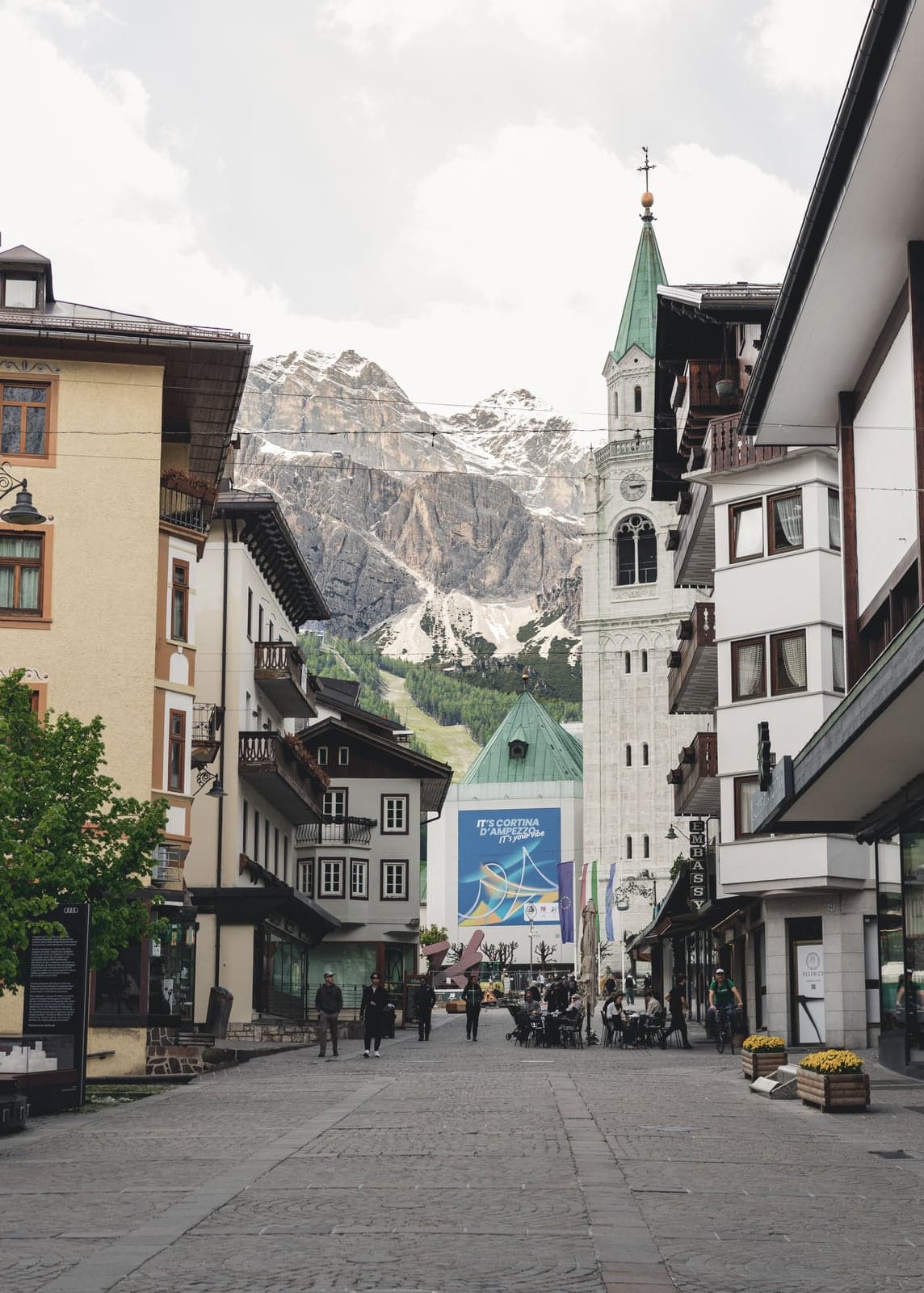 A cobbled pedestrian street in Cortina's center, with the church bell tower on the right and a snow-dusted Dolomite peak beyond
