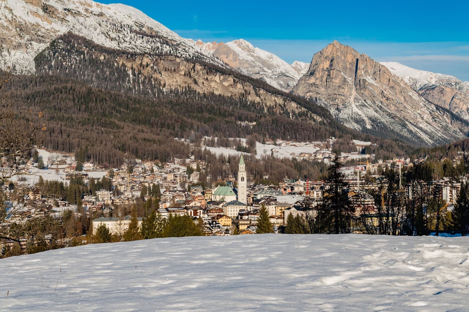 Cortina d'Ampezzo seen from a snowy slope above town, with the church bell tower among the houses and Dolomite peaks rising behind