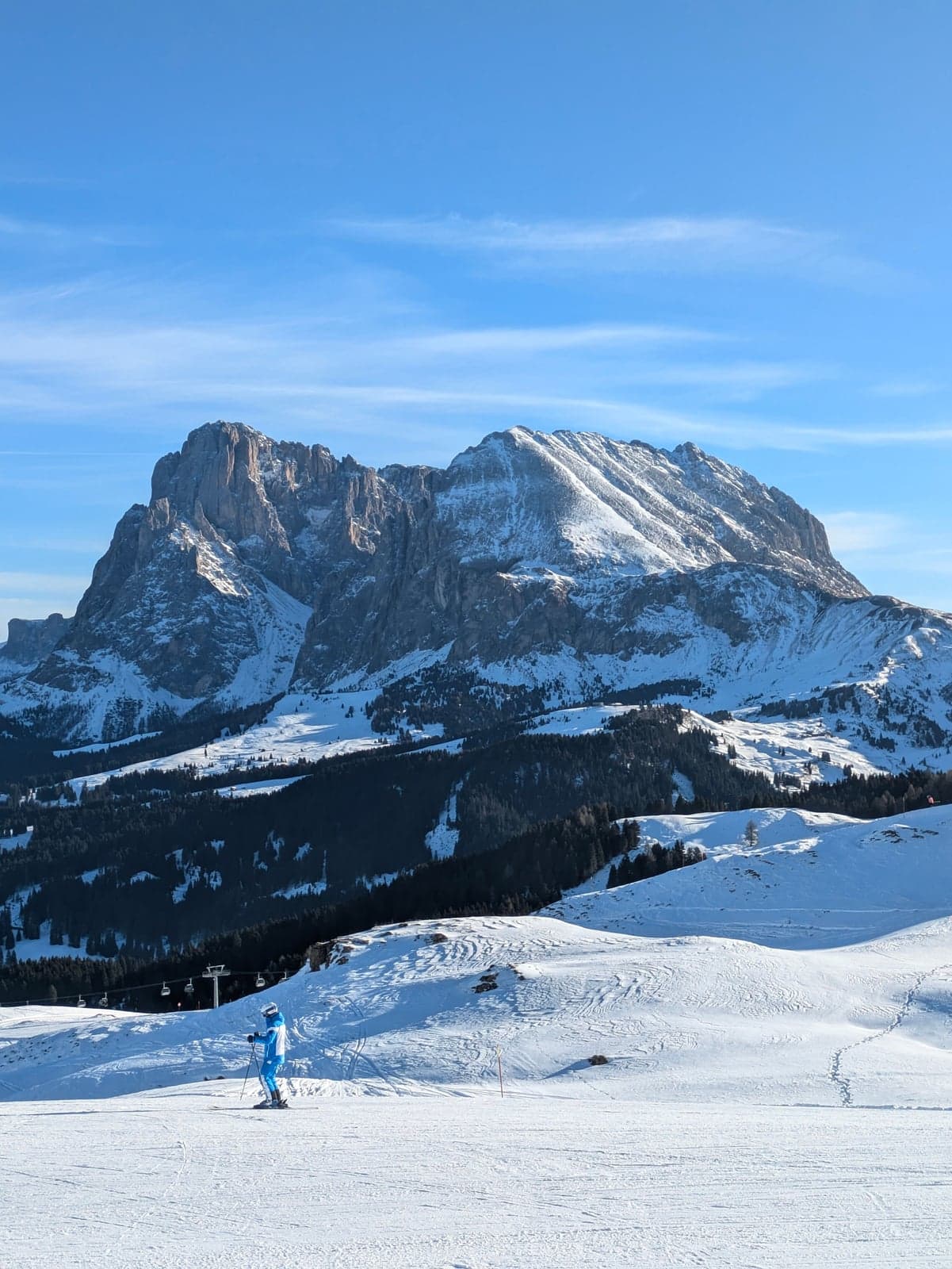Skier in a blue suit on a wide, gentle groomed slope with a ski lift line, and a rugged Dolomite massif rising behind under a clear sky