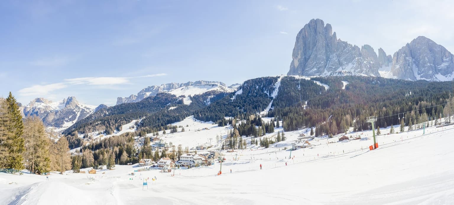 Wide view of gentle beginner slopes with a small lift and a few skiers, village buildings at the base and the Sassolungo rock wall on the right