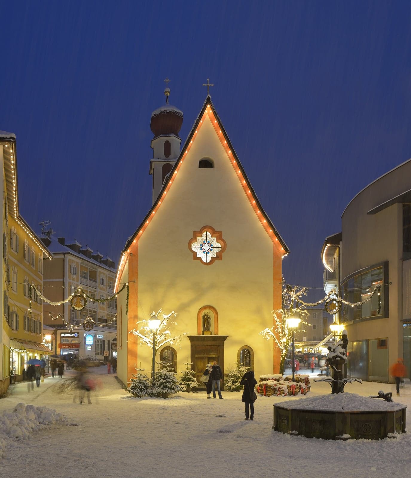 Small chapel outlined in warm lights with the onion-domed church tower behind it on a snowy square in Ortisei at dusk, with Christmas decorations and a fountain