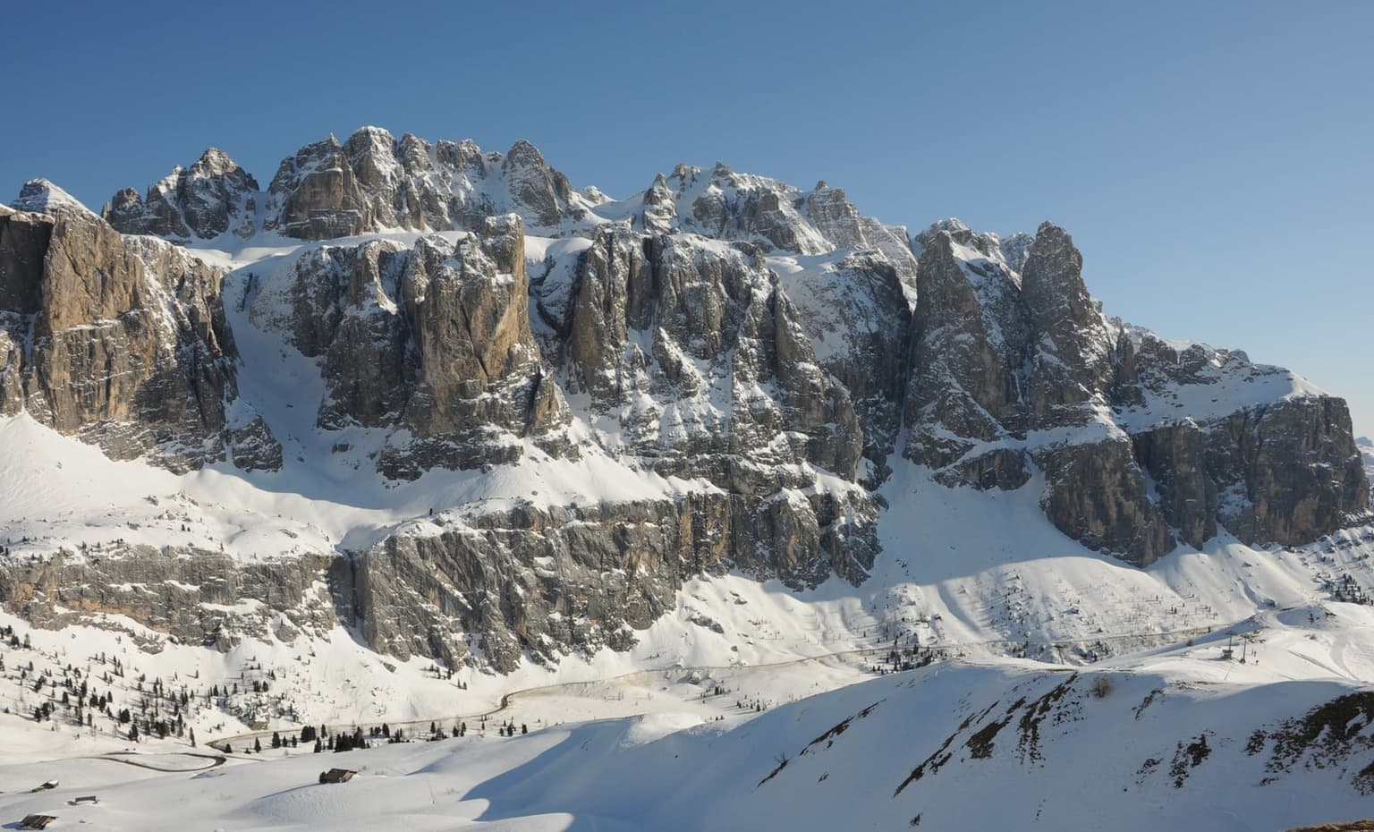 Sunlit rock towers of the Sella massif above the snow-covered Passo Gardena saddle, with a road and lift station below