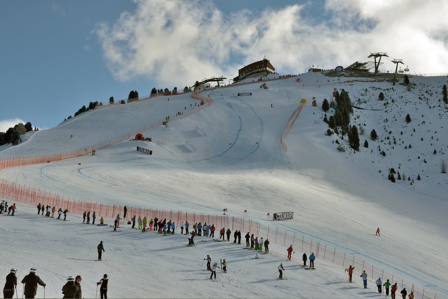 Saslong World Cup downhill course with orange safety netting and sponsor boards, skiers and spectators along the edge and a lift station at the top of the slope