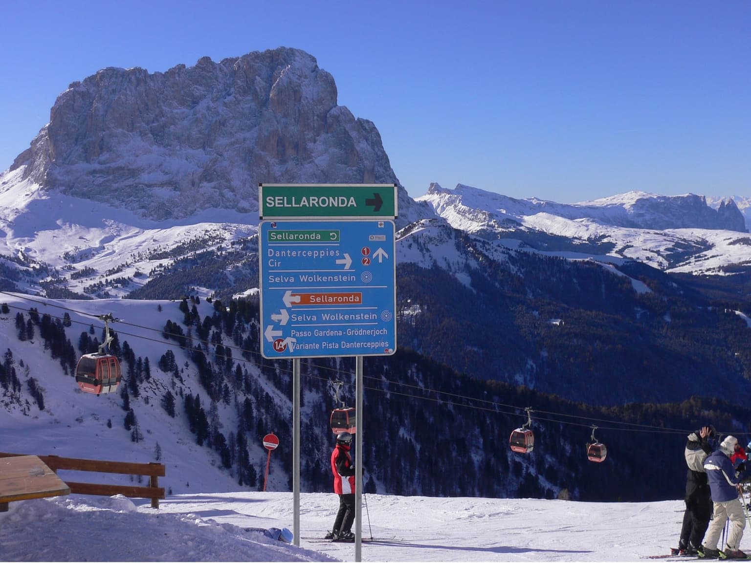 Sellaronda signpost at a lift and piste junction, with orange and green direction signs, gondola cabins and the Sassolungo massif behind