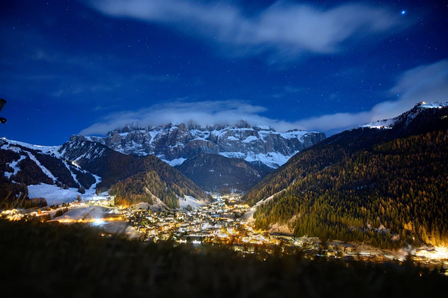 Village lights glowing at night in a snowy Dolomite valley, with a ski slope on the left hillside and the snow-covered Sella massif behind
