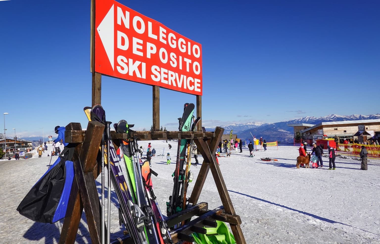 Ski rental and storage point with a wooden rack of skis and boots under a red Noleggio Deposito Ski Service sign, with a busy snowy base area behind