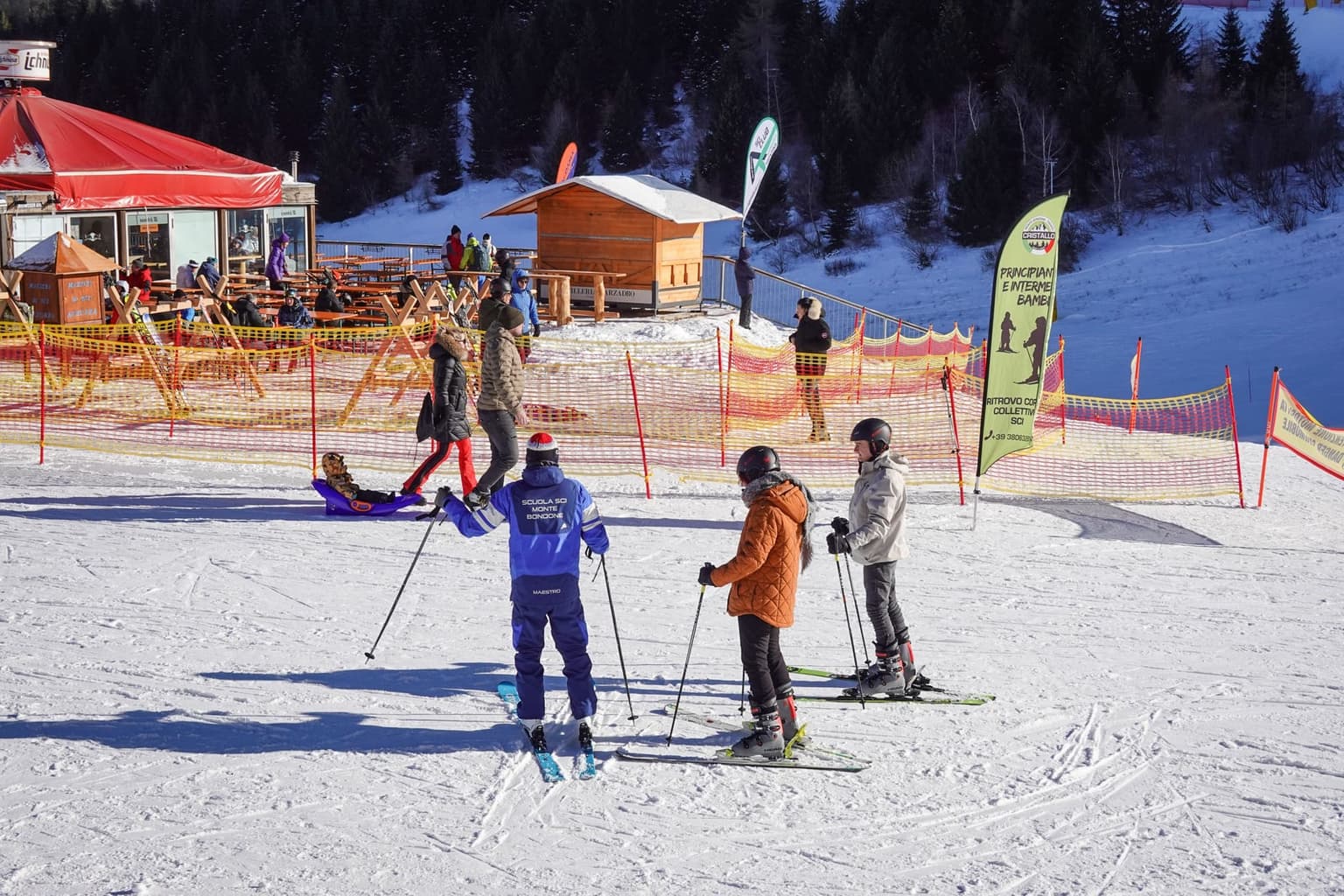 Ski instructor in a blue school jacket facing a small group of adult beginners on a gentle snowy slope beside a mountain lodge