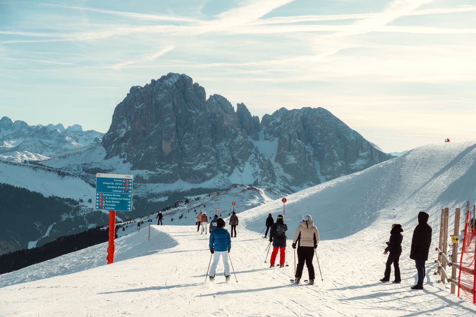 Skiers heading down a groomed piste toward the Sassolungo rock wall, with a signpost to Ortisei, Gardena Ronda and Sella Ronda beside the run