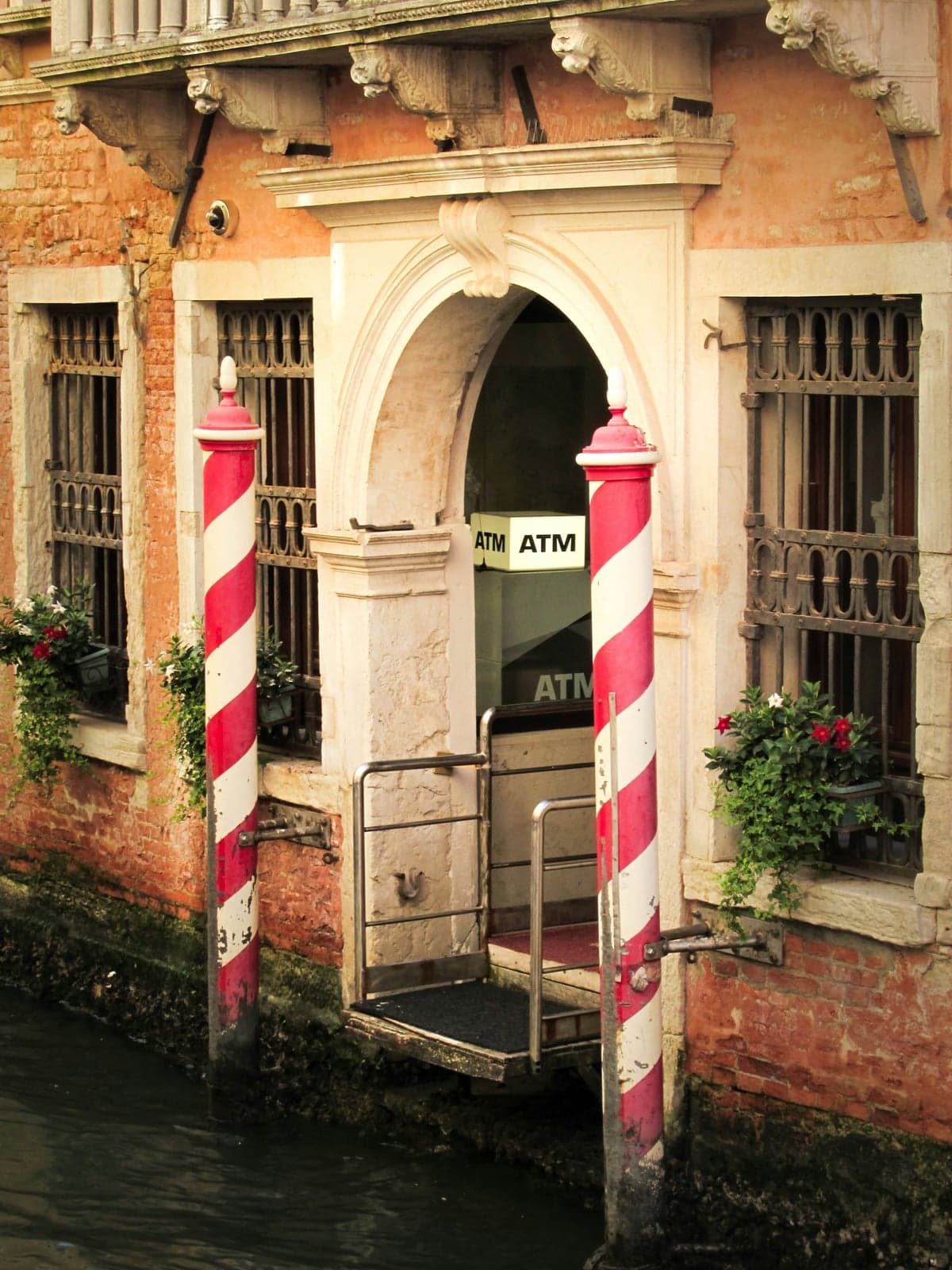 A cash machine with a lit ATM sign inside an arched canal-side doorway in Venice, framed by red-and-white striped mooring poles