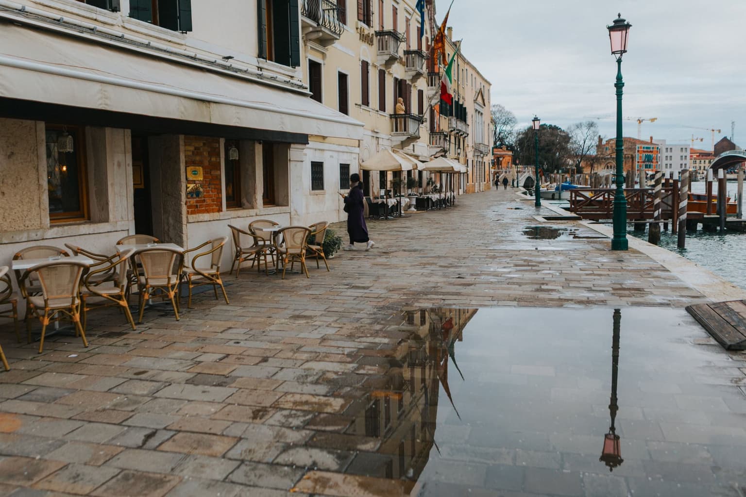 A wet stone quay along a Venice canal on a gray day, a large puddle reflecting a street lamp beside empty cafe chairs