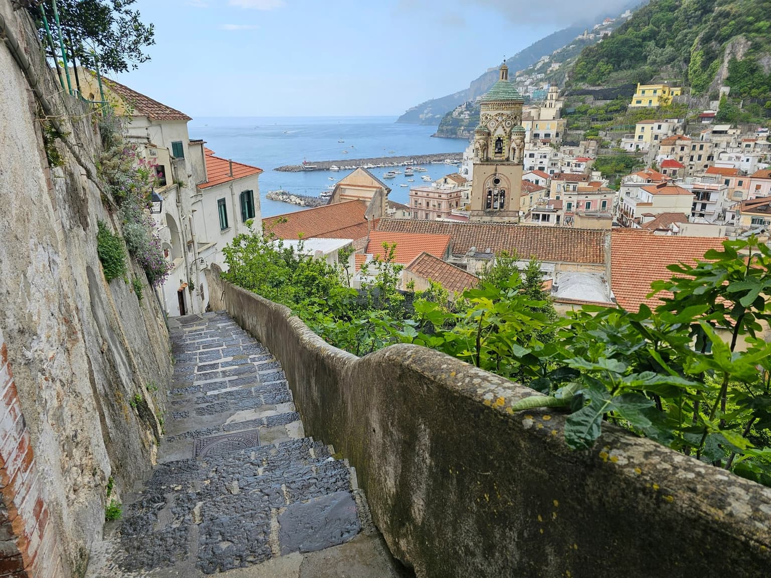 Worn stone steps drop steeply between a wall and a low parapet toward Amalfi's red roofs, cathedral bell tower, and the sea
