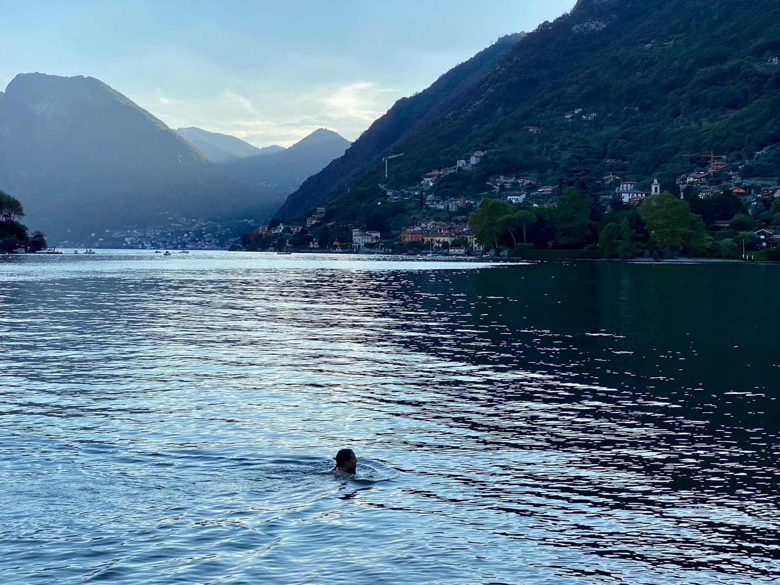 A lone swimmer in the open water of Lake Como at dusk, a village and mountains behind