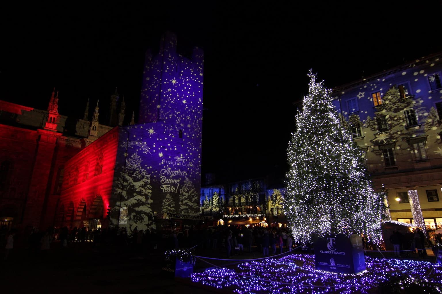Como's Città dei Balocchi festival at night: a lit Christmas tree and colored light projections on the medieval tower, crowds on the square