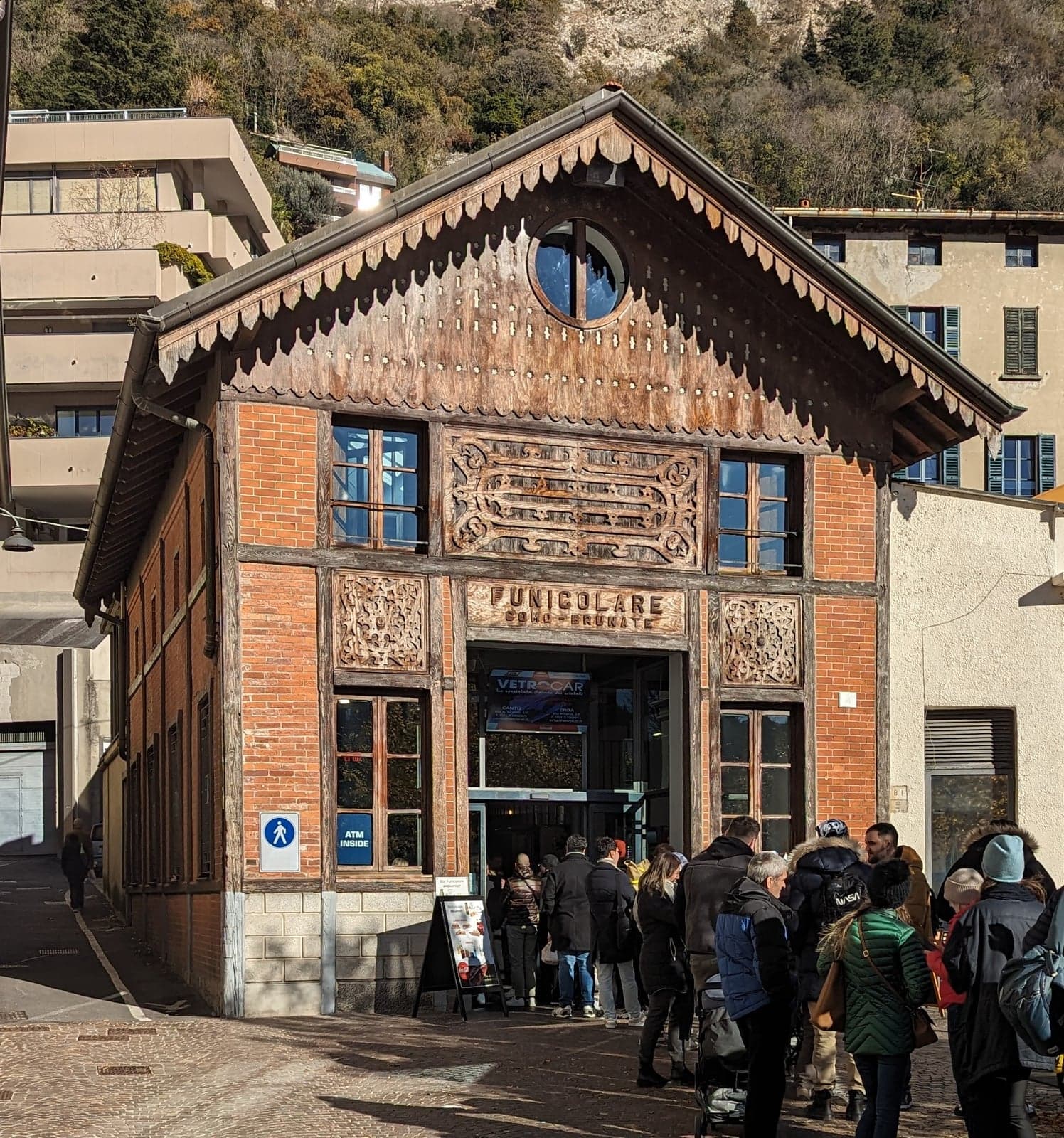 The carved wooden facade of the Como-Brunate funicular's lower station, its "Funicolare Como-Brunate" sign above the entrance, directly opposite the Como Nord Lago exit