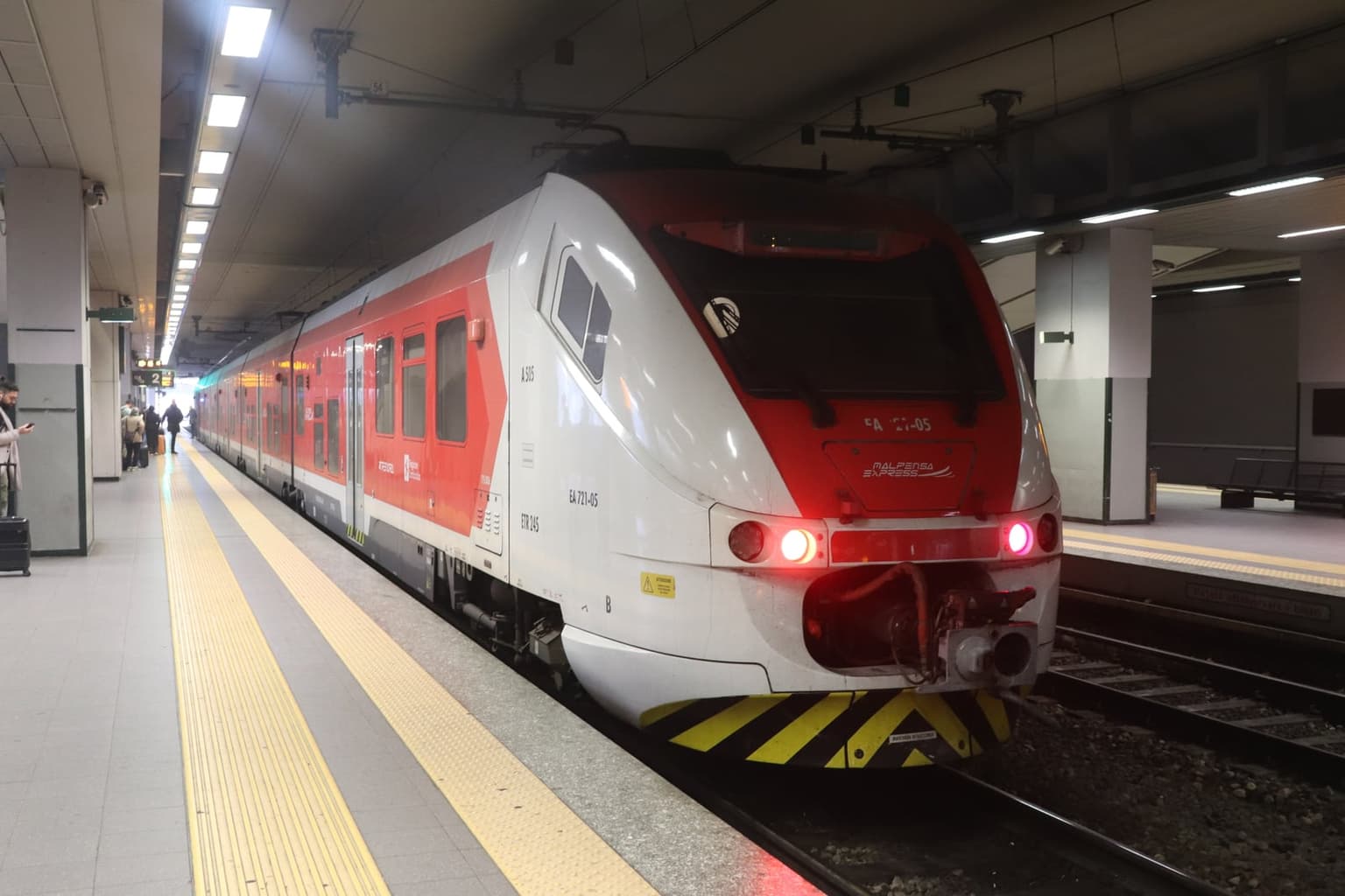 A red-and-white Malpensa Express train at the platform inside Malpensa Aeroporto T1 station, the airport's rail link into Milan and onward to the lake