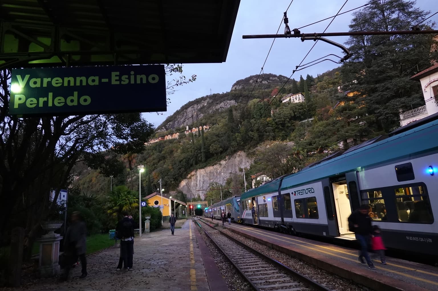 Arriving at Varenna-Esino Perledo station at dusk, a Trenord regional train at the platform below the wooded hillside