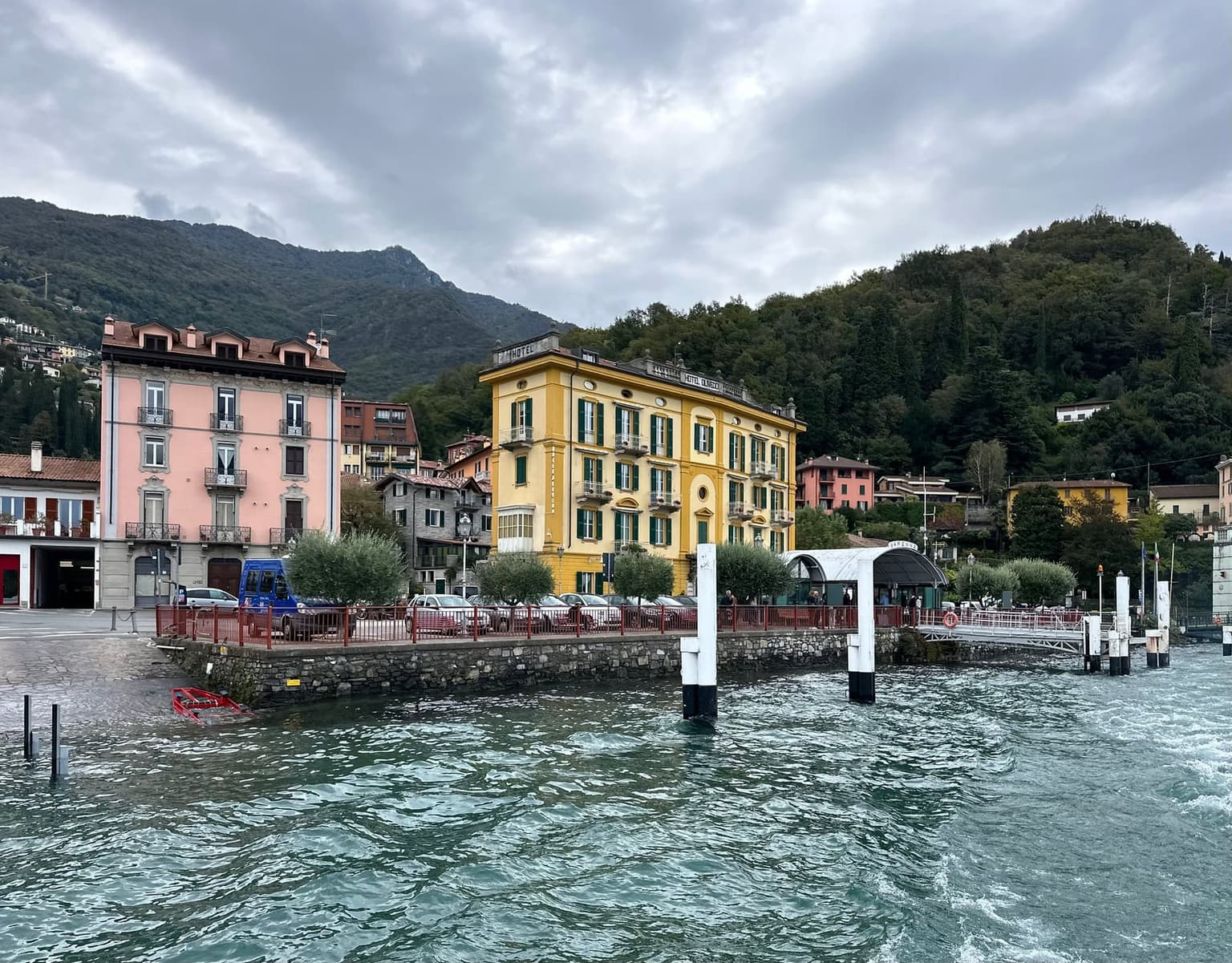 The Varenna ferry landing with its covered imbarcadero and the yellow Hotel Olivedo behind, cars parked along the waterfront on an overcast day