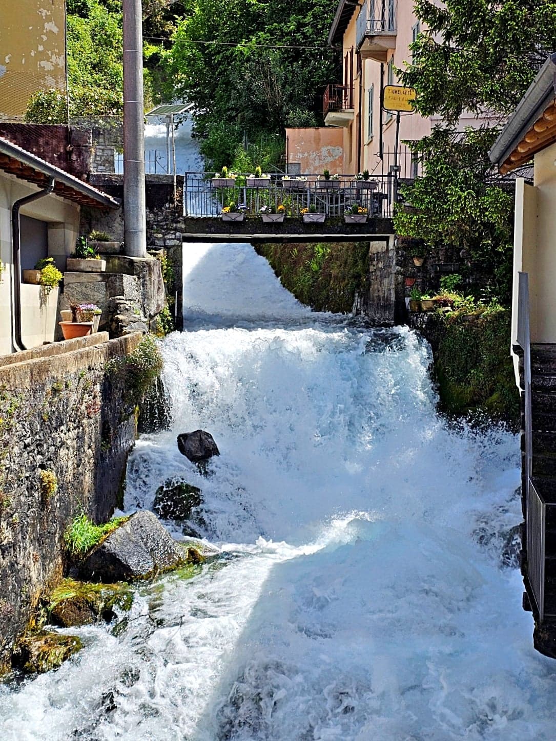 The milky-white Fiumelatte torrent foaming down between village houses near Varenna, its yellow name sign on the wall