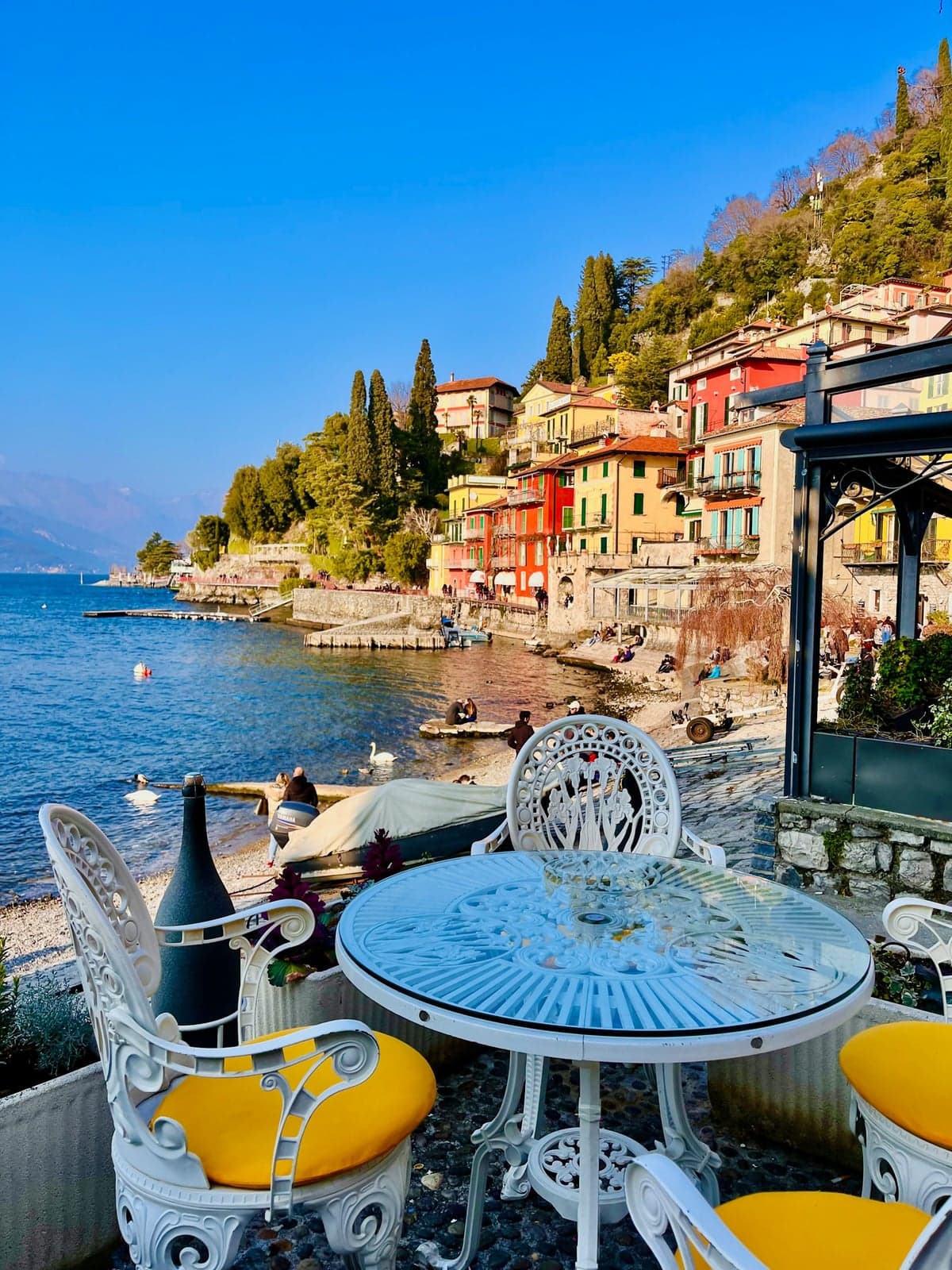 A white cafe table with yellow cushions on a Varenna waterfront terrace, the village's colourful lakeside houses and the lake beyond