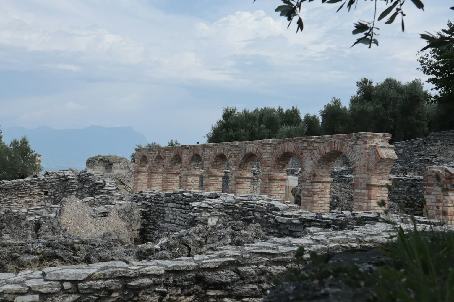 The Roman ruins of the Grotte di Catullo in Sirmione, a row of brick arches above layered stone foundations among olive trees, with a hazy mountain ridge in the distance