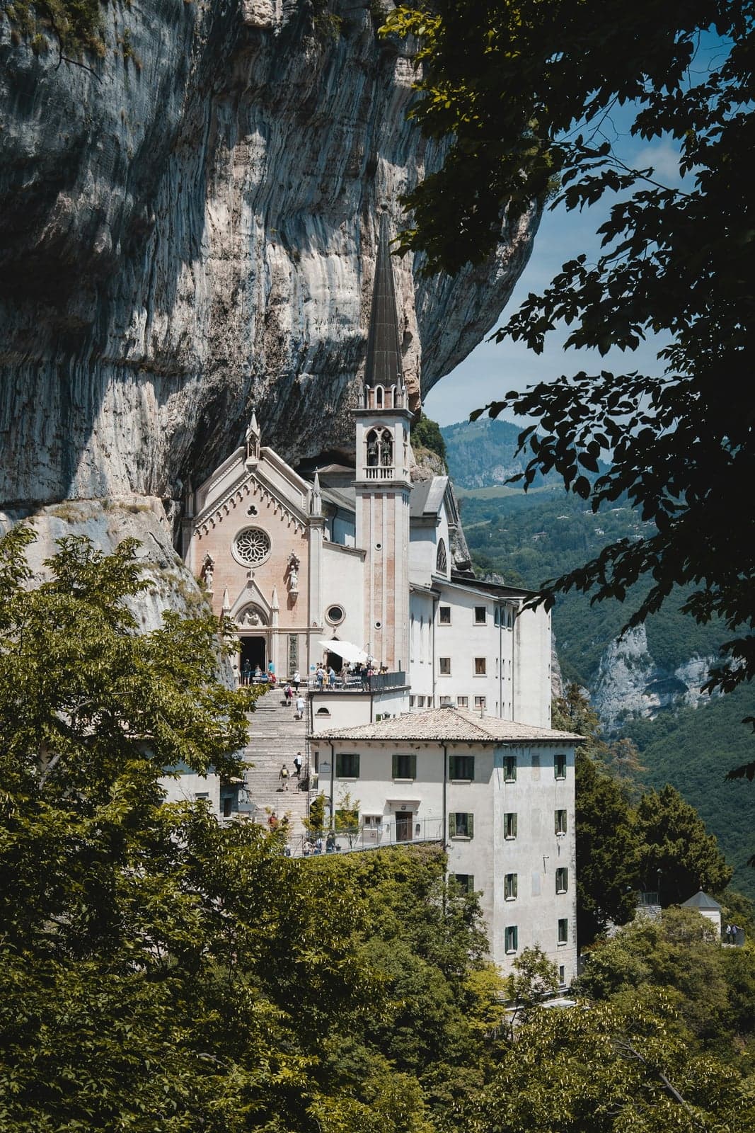 The sanctuary of Madonna della Corona built into a sheer cliff face, its pale facade and slender bell tower above a steep stairway with visitors, a wooded valley dropping away to the right