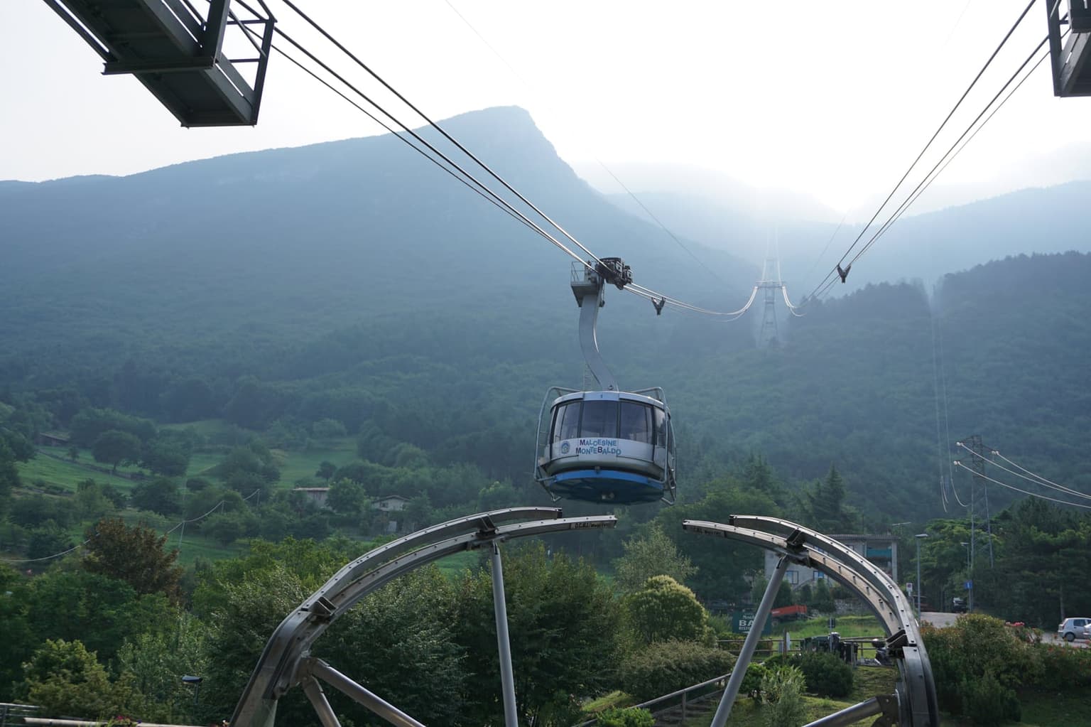 A rounded glass cabin of the Malcesine to Monte Baldo cable car leaving the station on its cables, with a pylon and a hazy forested mountainside behind