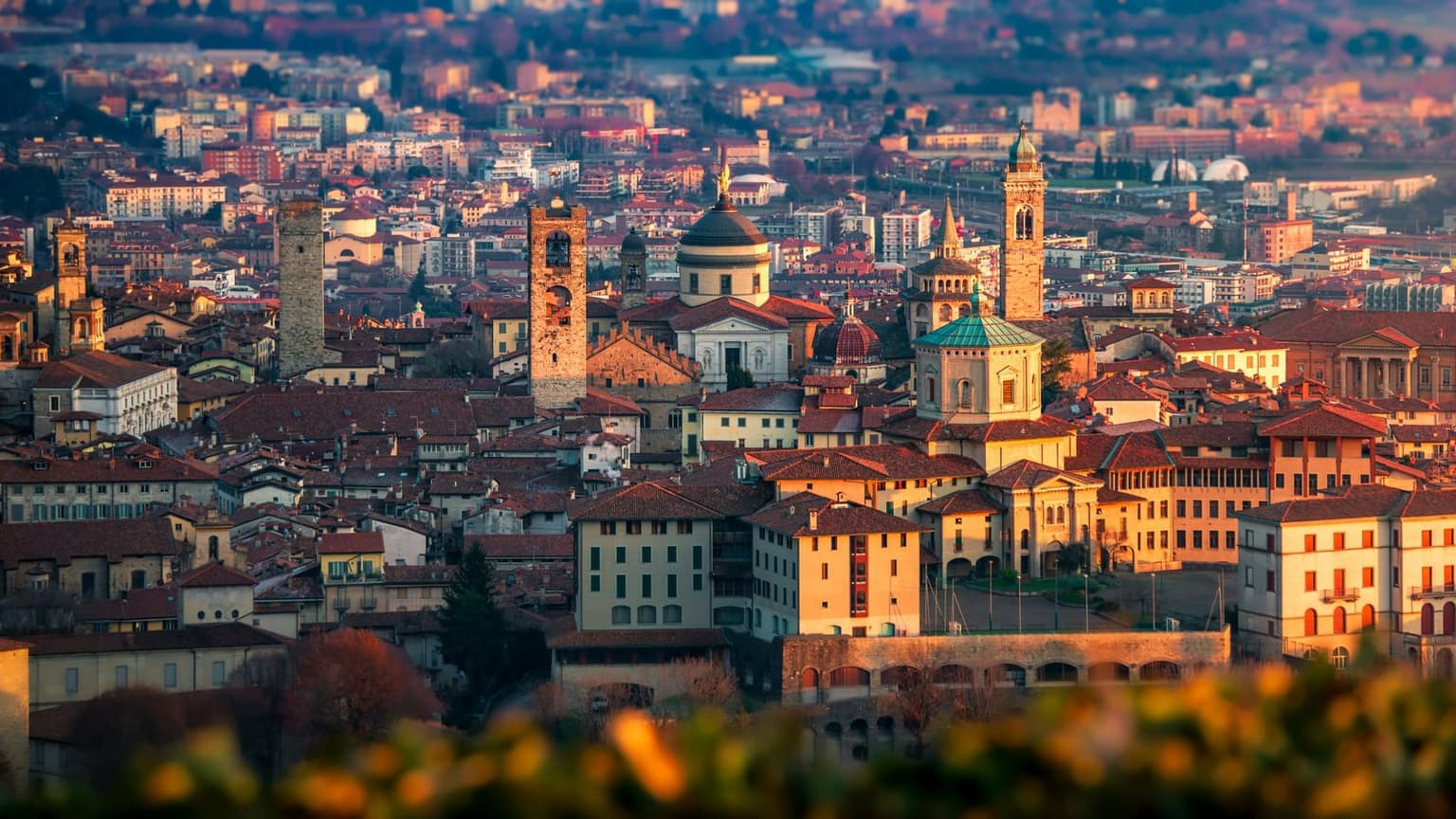 Bergamo's upper town from above at golden hour, medieval towers, domes and bell towers over terracotta roofs, with the lower city fading into the haze behind