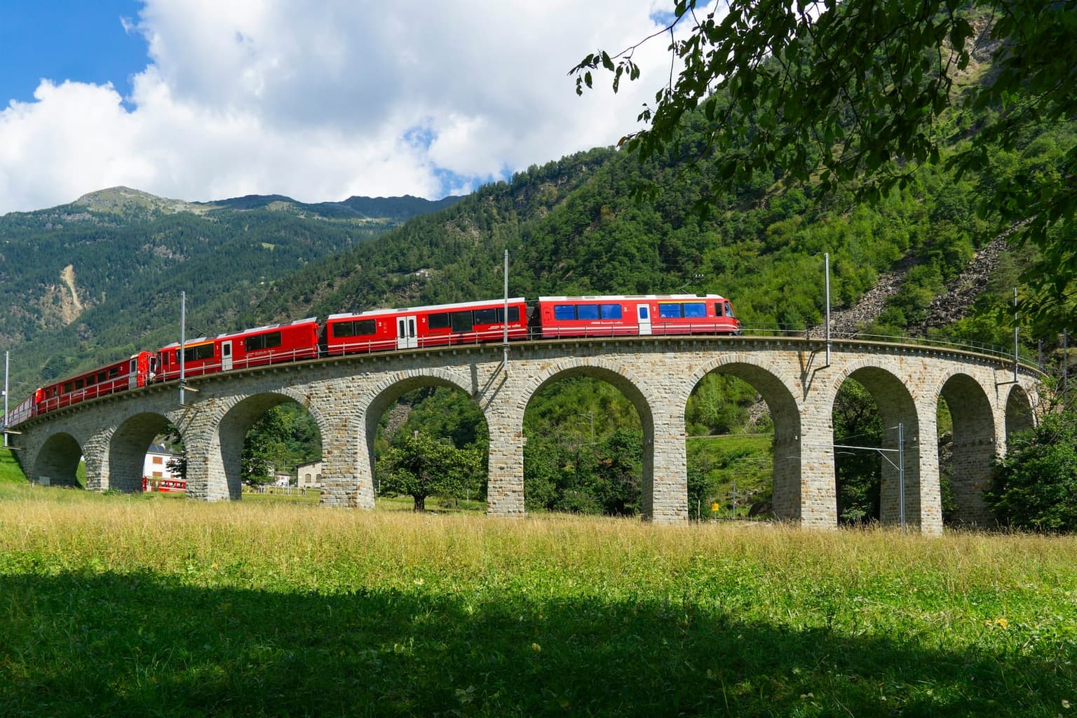A red train crossing the curving stone arches of the Brusio circular viaduct, a sunny meadow in front and wooded mountains behind