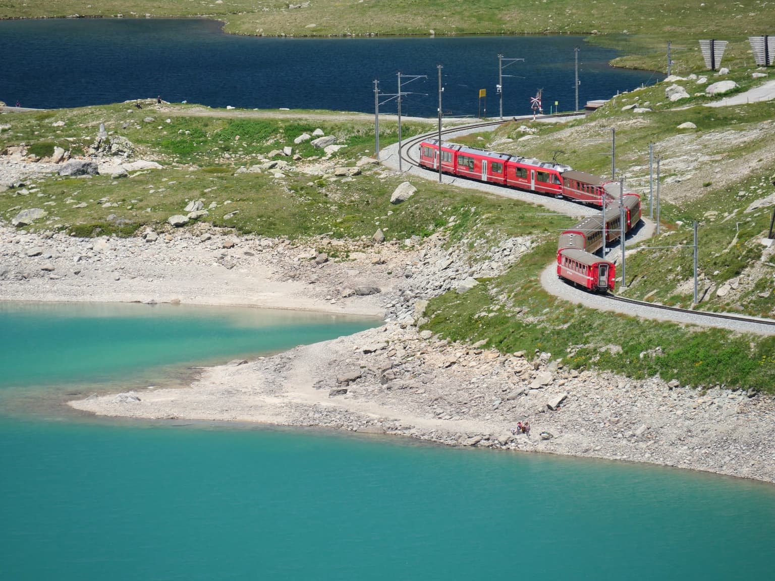 A red train curving along the shore of a turquoise high-alpine lake on the Bernina Pass, rocky ground all around and a darker lake behind