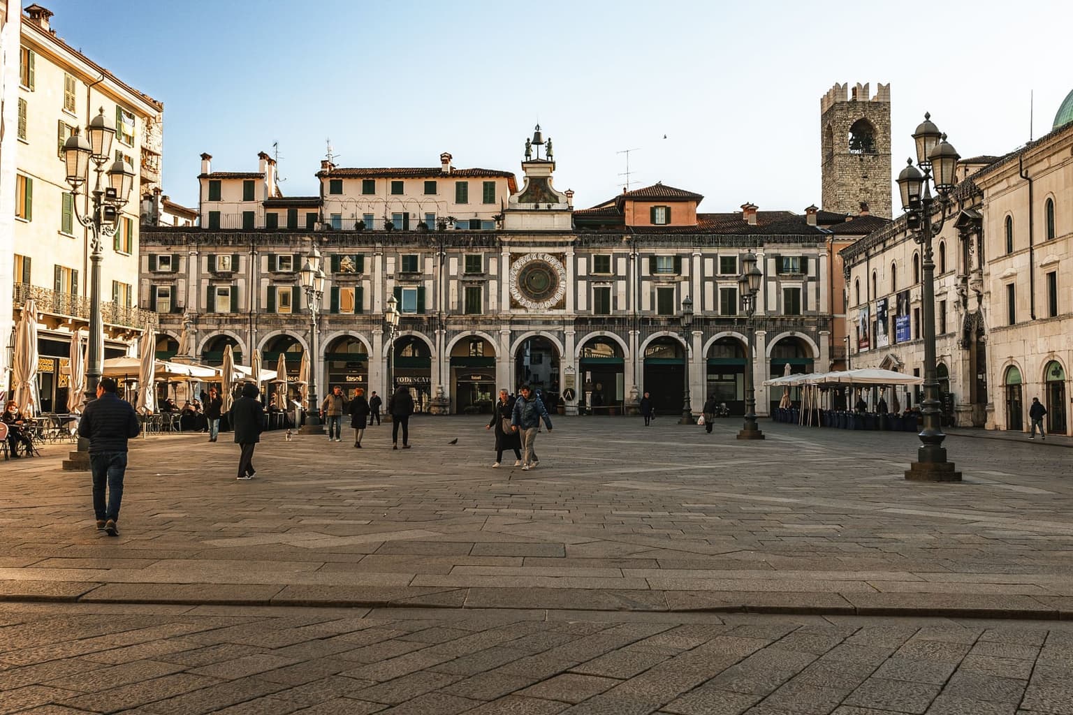 Brescia's Piazza della Loggia, an arcaded facade with a large astronomical clock and two bell-ringer figures facing the paved square, a crenellated stone tower behind