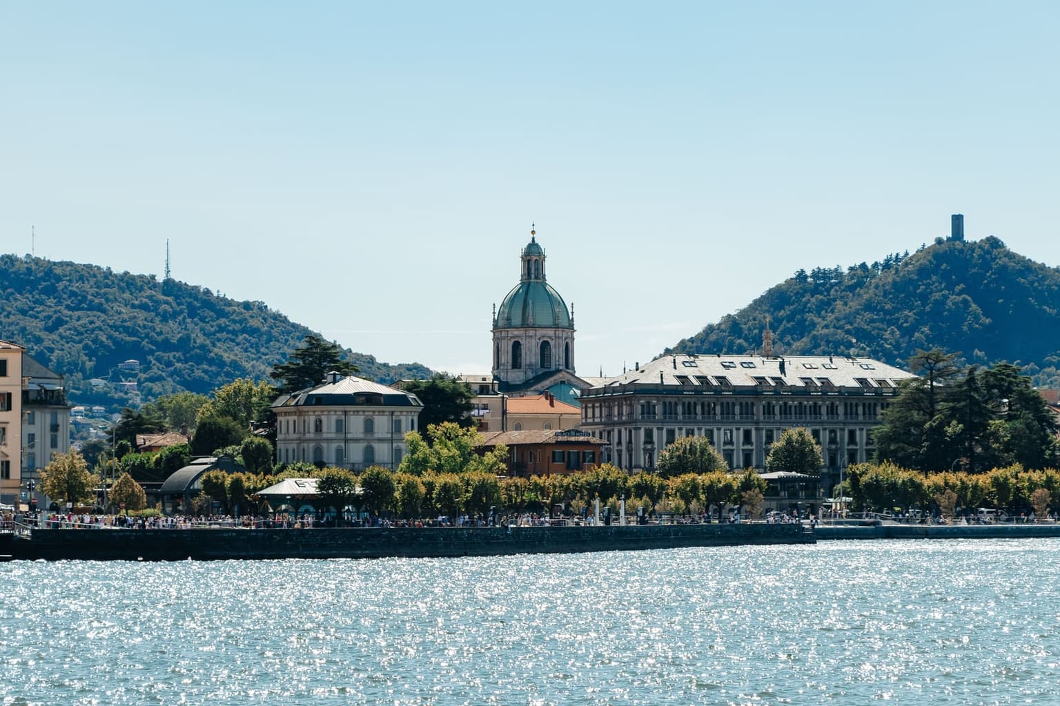 Como from the lake, the cathedral's green dome above the waterfront buildings and a tree-lined promenade, with wooded hills behind and sun glinting on the water